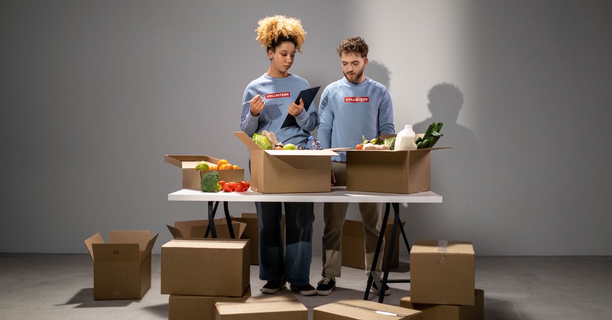 Two volunteers organizing food donations in cardboard boxes for charity indoors.